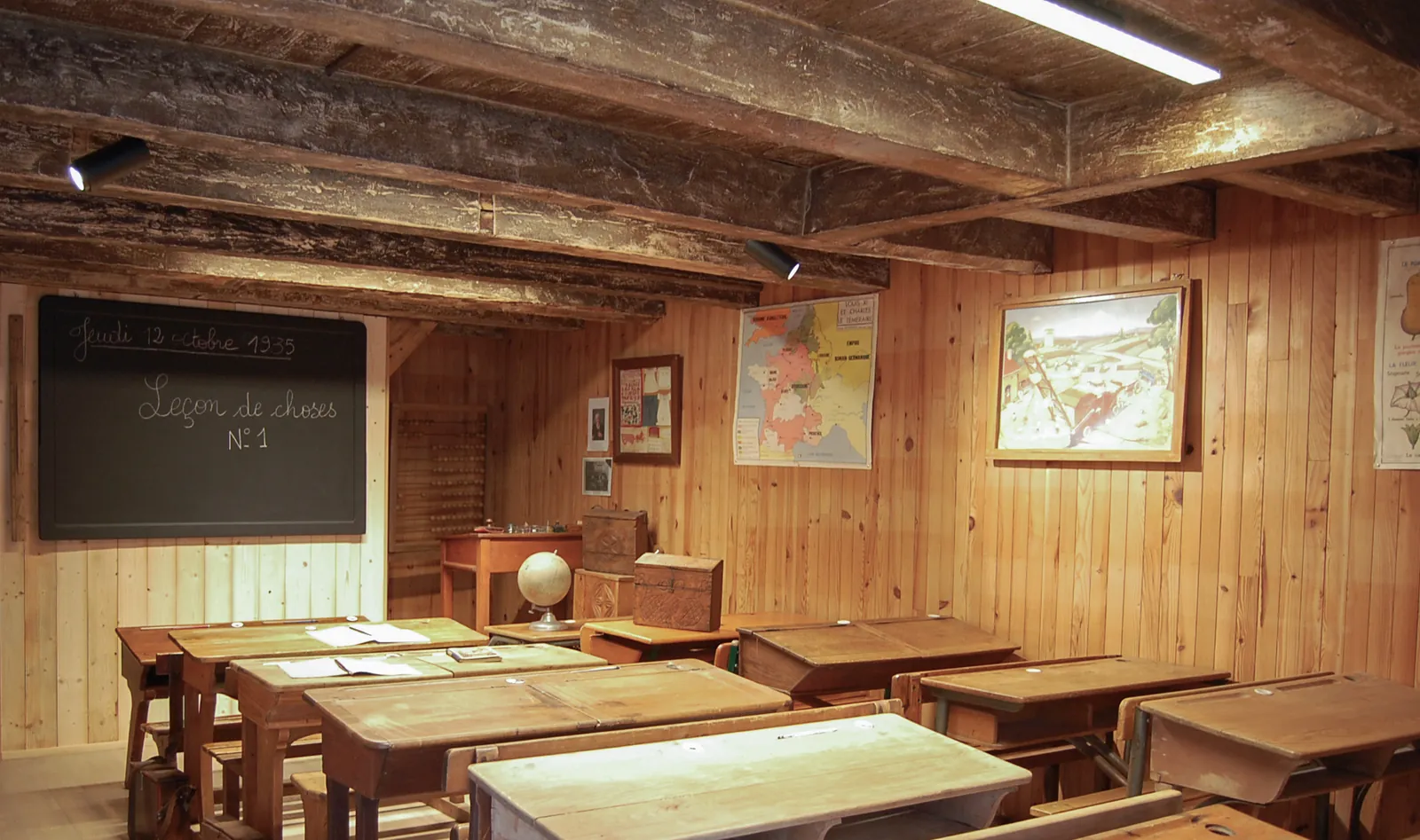 Salle de classe d'autrefois reconstituée au musée de Fessy : pupitres en bois alignés, tableau noir portant l'inscription « Jeudi 12 octobre 1935, Leçon de choses n° 1 », globe terrestre et cartes murales.